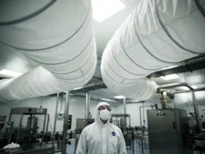 Farmaceutische productieruimte met witte luchtfiltersokken aan plafond, technicus in beschermende kleding inspecteert systeem