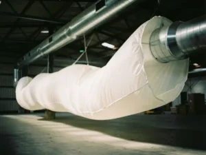 Witte stoffen luchtkanaal hangt aan industriële balken in magazijn, zacht bollend door luchtstroom met dramatische belichting